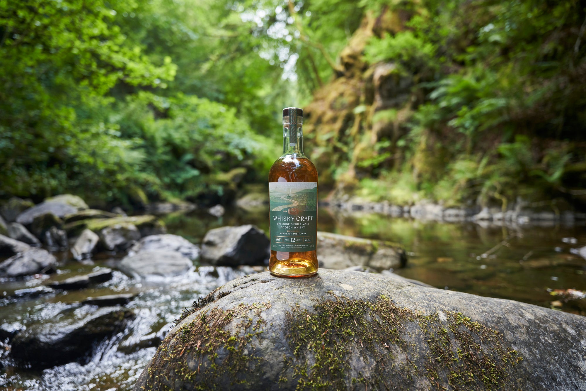 Bottle of Mortlach whisky on a rock next to a small stream in with steep and lush forested banks