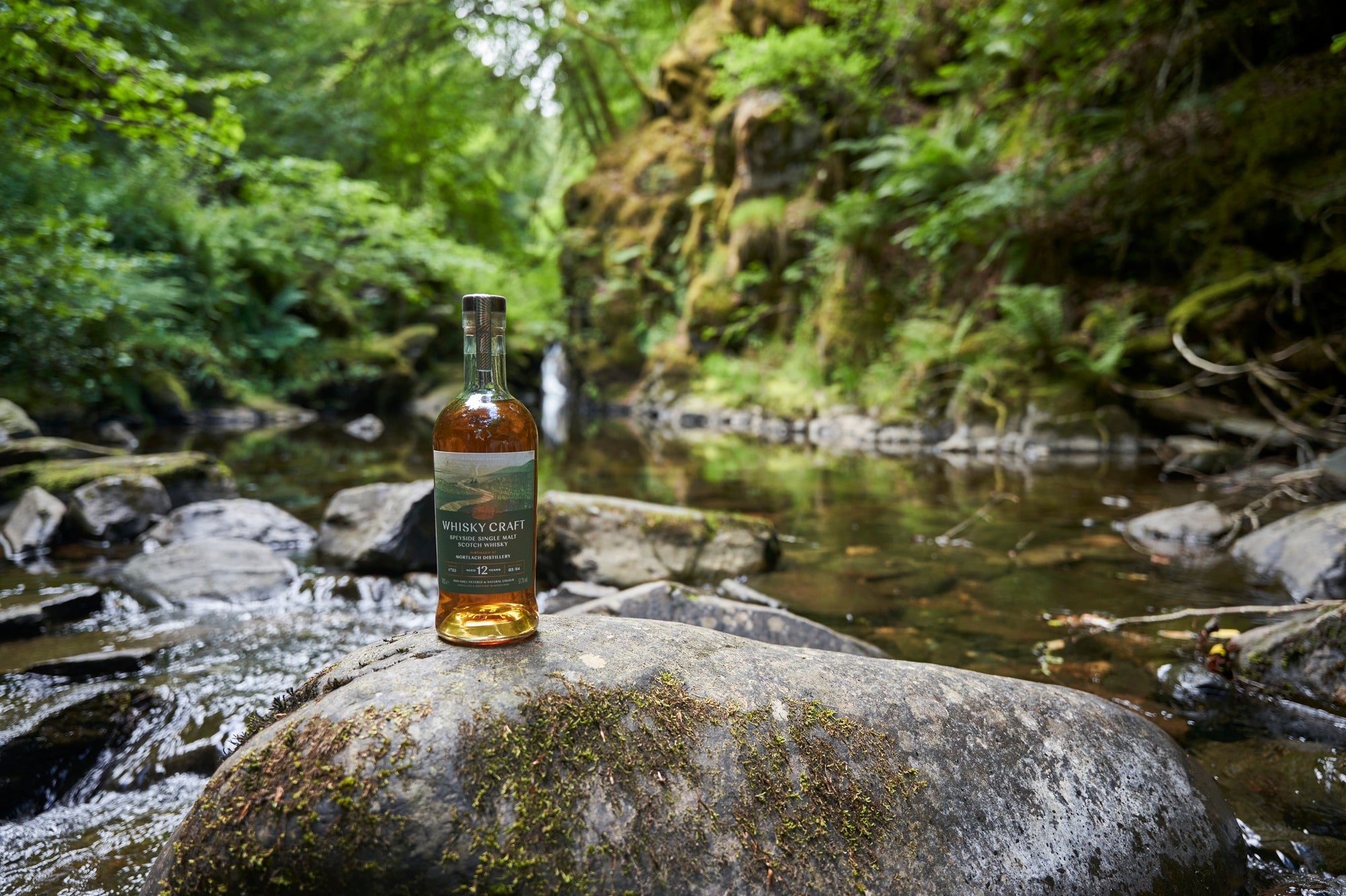 Bottle of Mortlach whisky on a rock next to a small stream in with steep and lush forested banks