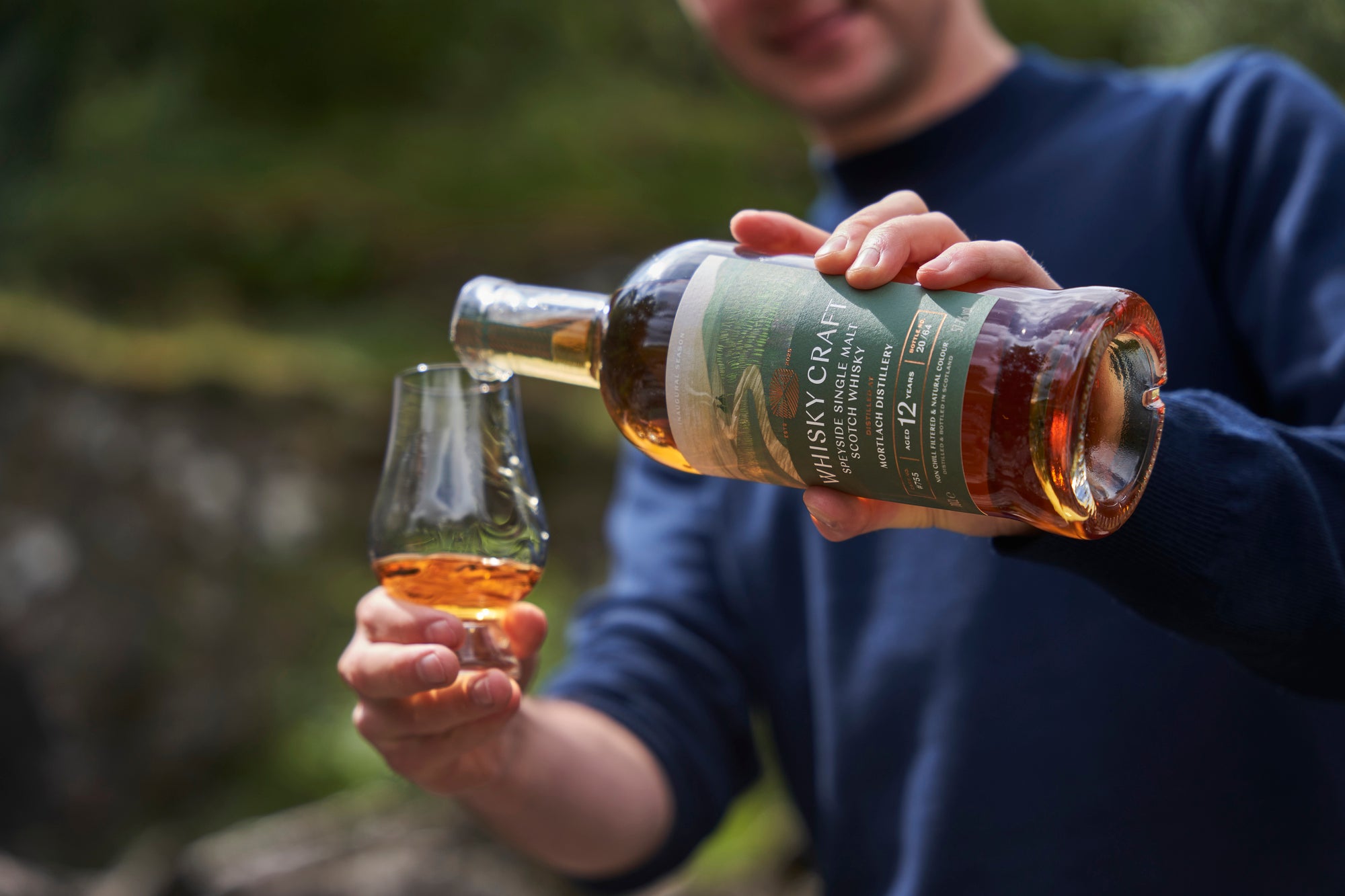 Someone pouring a bottle of Mortlach whisky into a glass, in front of an out-of-focus natural background