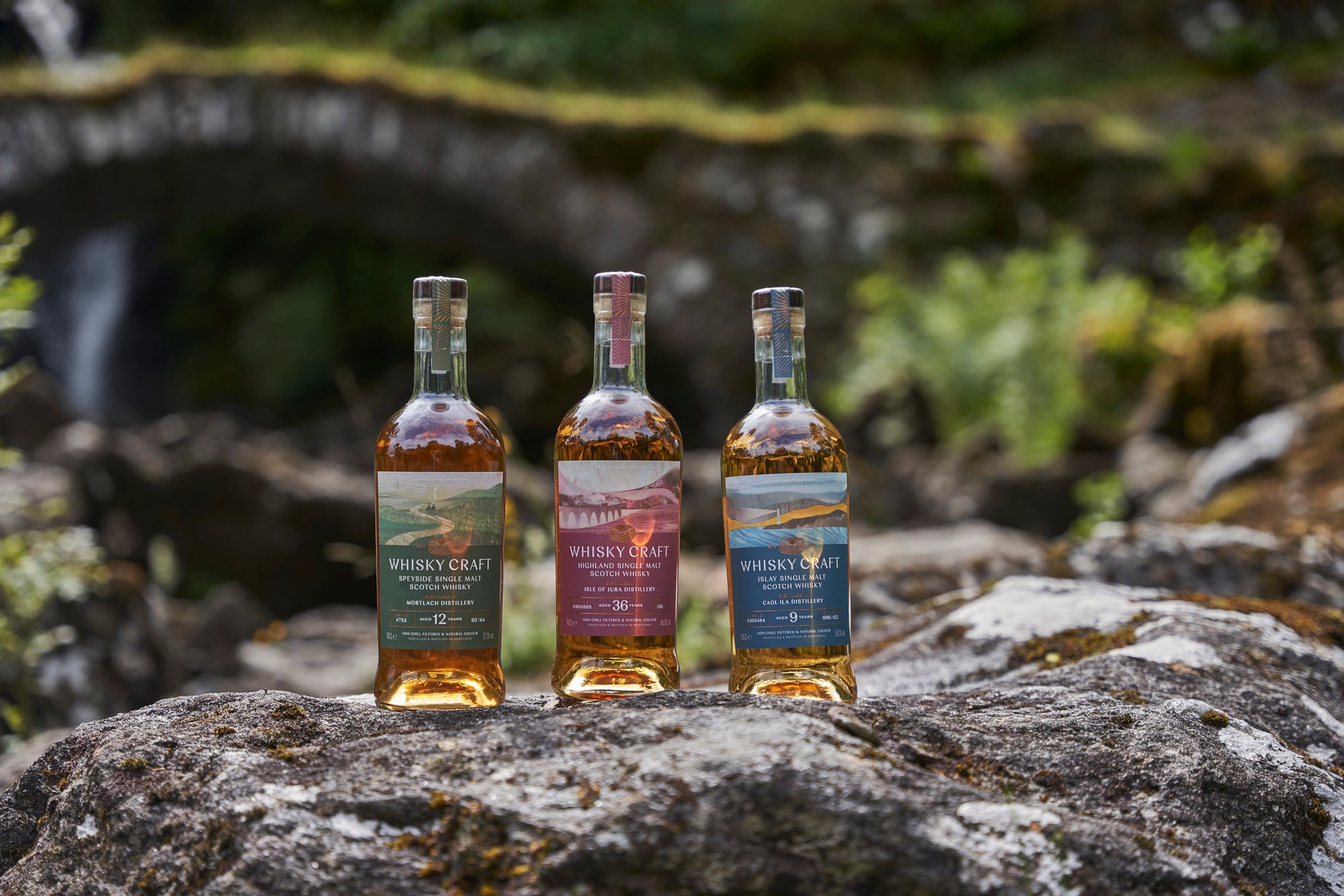 Close-up of three bottles of whisky on a rock in front of an old stone bridge. It looks very wild and lush