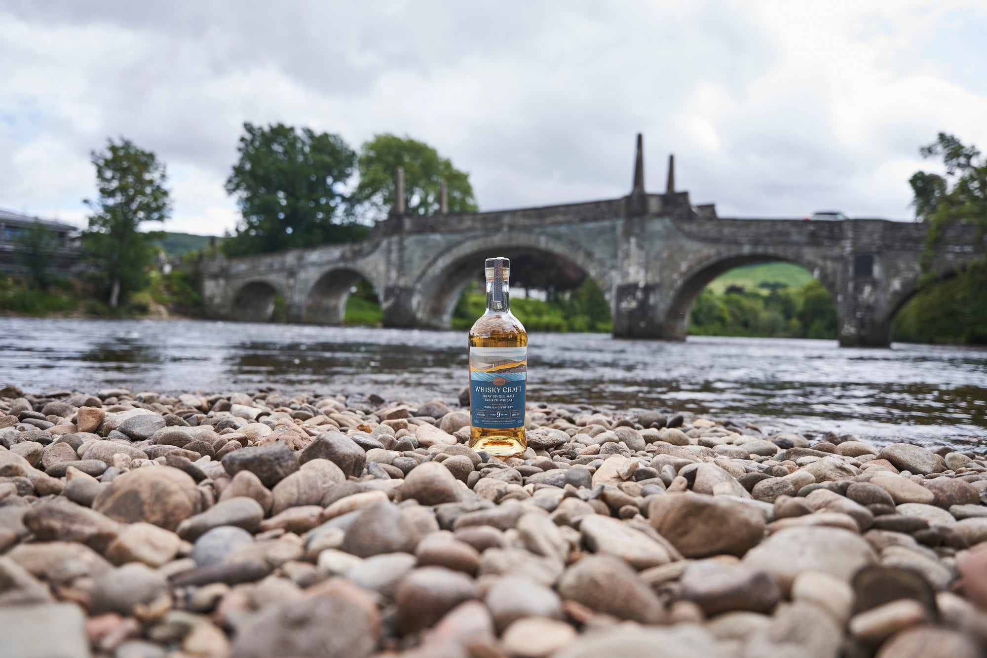 Bottle of Islay whisky on a rock beach in front of a large river an big, multi-arched gothic stone bridge