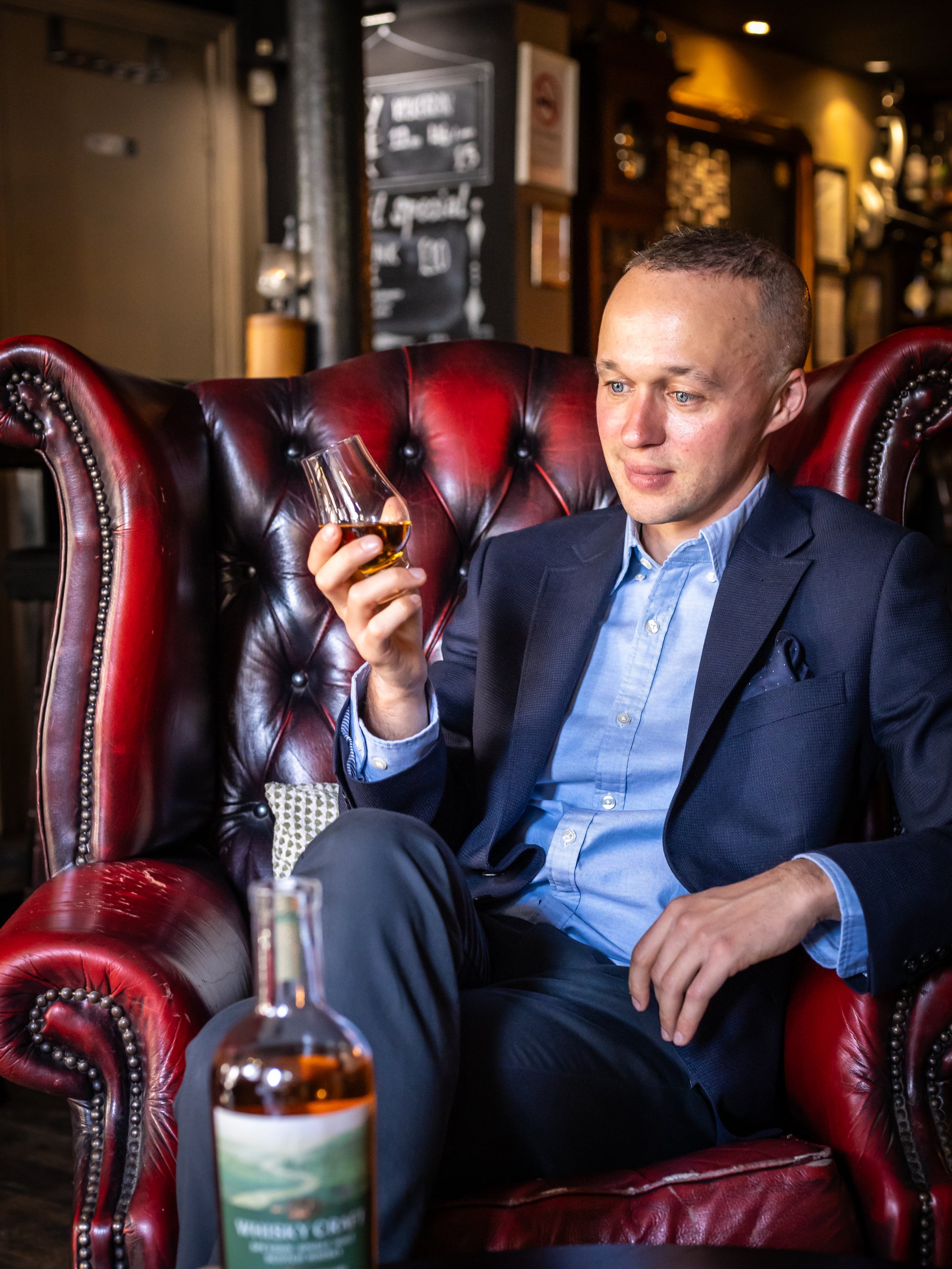 Sonny sitting in a large, ornate red armchair, looking at a dram of whisky in his hand. He's wearing a suit jacket and blue shirt, the bottle is in the foreground, and the background is very decorated.